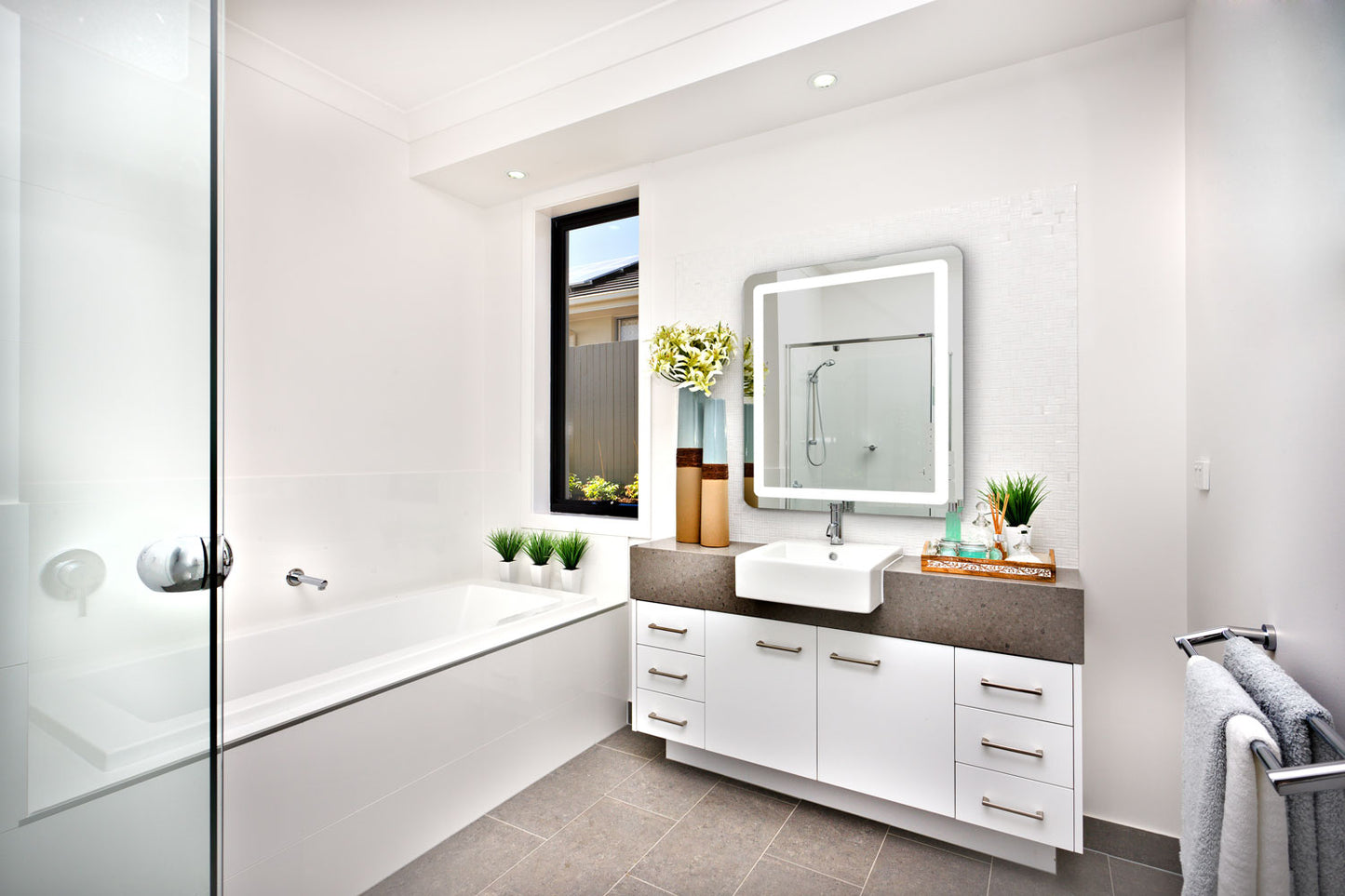 A bright modern bathroom featuring a bathtub, a vanity with a rounded rectangle lighted mirror, and decorative plants arranged throughout the space.