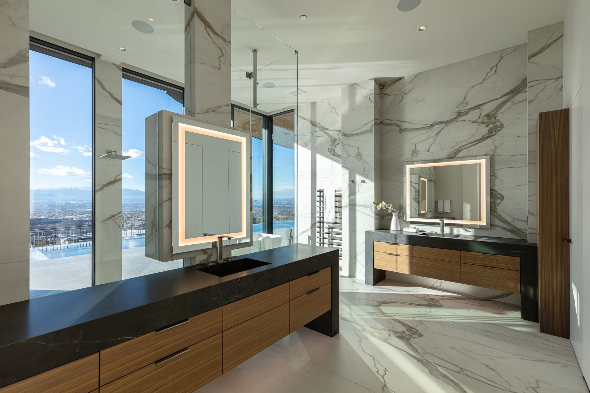 Modern bathroom with marble walls, wood vanities, black countertops, and lighted mirrors with natural light.

