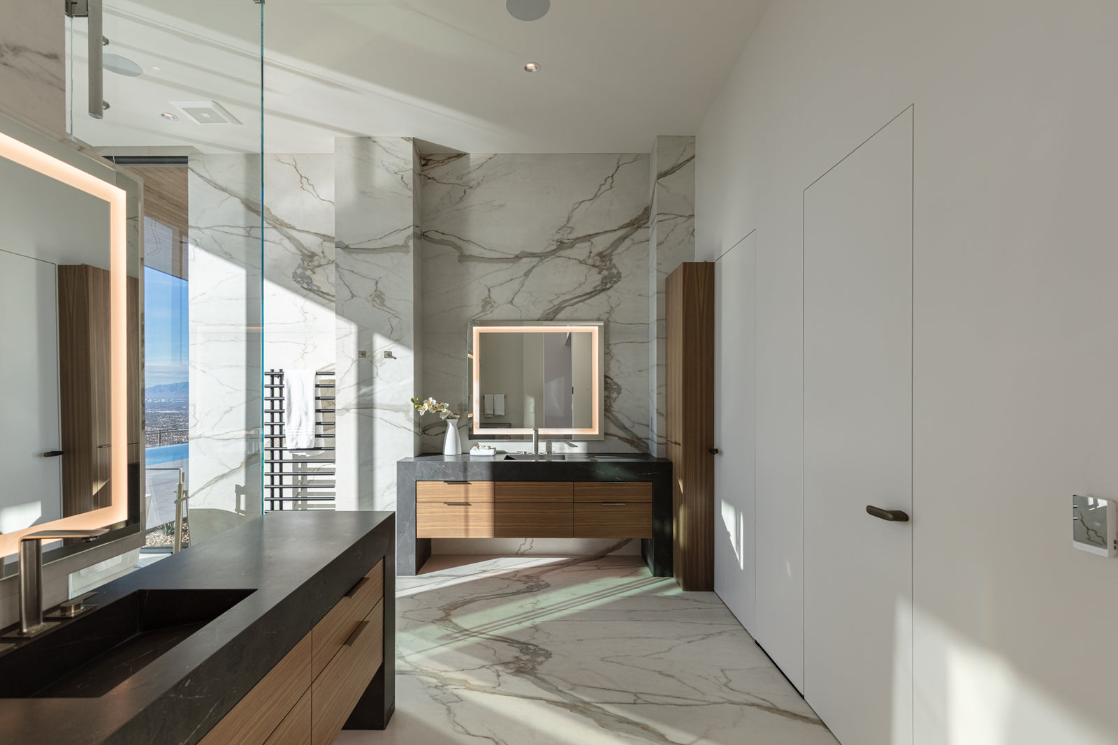 Modern bathroom with marble walls and floors, a wood vanity, black countertop, and an illuminated mirror with natural light.