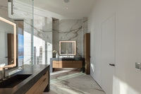Modern bathroom with marble walls and floors, a wood vanity, black countertop, and an illuminated mirror with natural light.