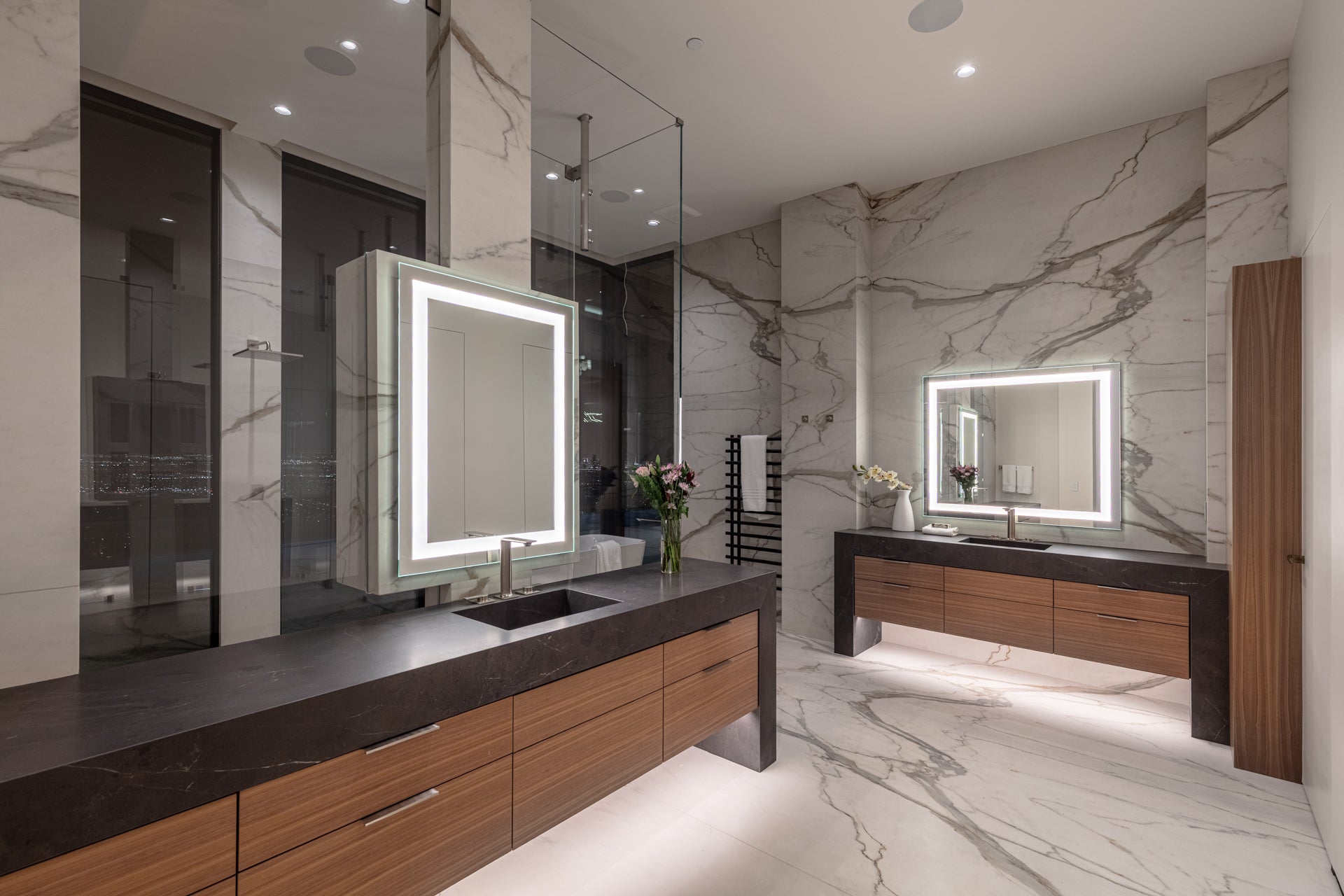 Modern bathroom with marble walls and floors, wood vanities, black countertops, and lighted mirrors.

