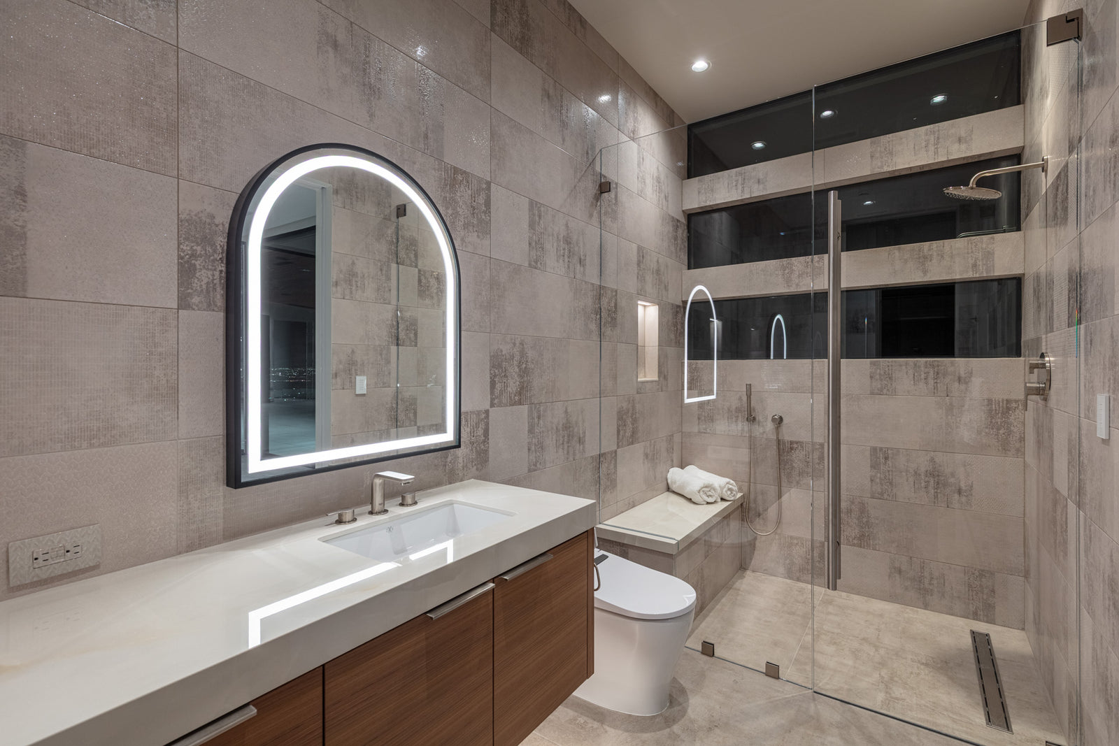 Modern bathroom with a wood vanity, illuminated mirror, wall-mounted toilet, and glass shower.

