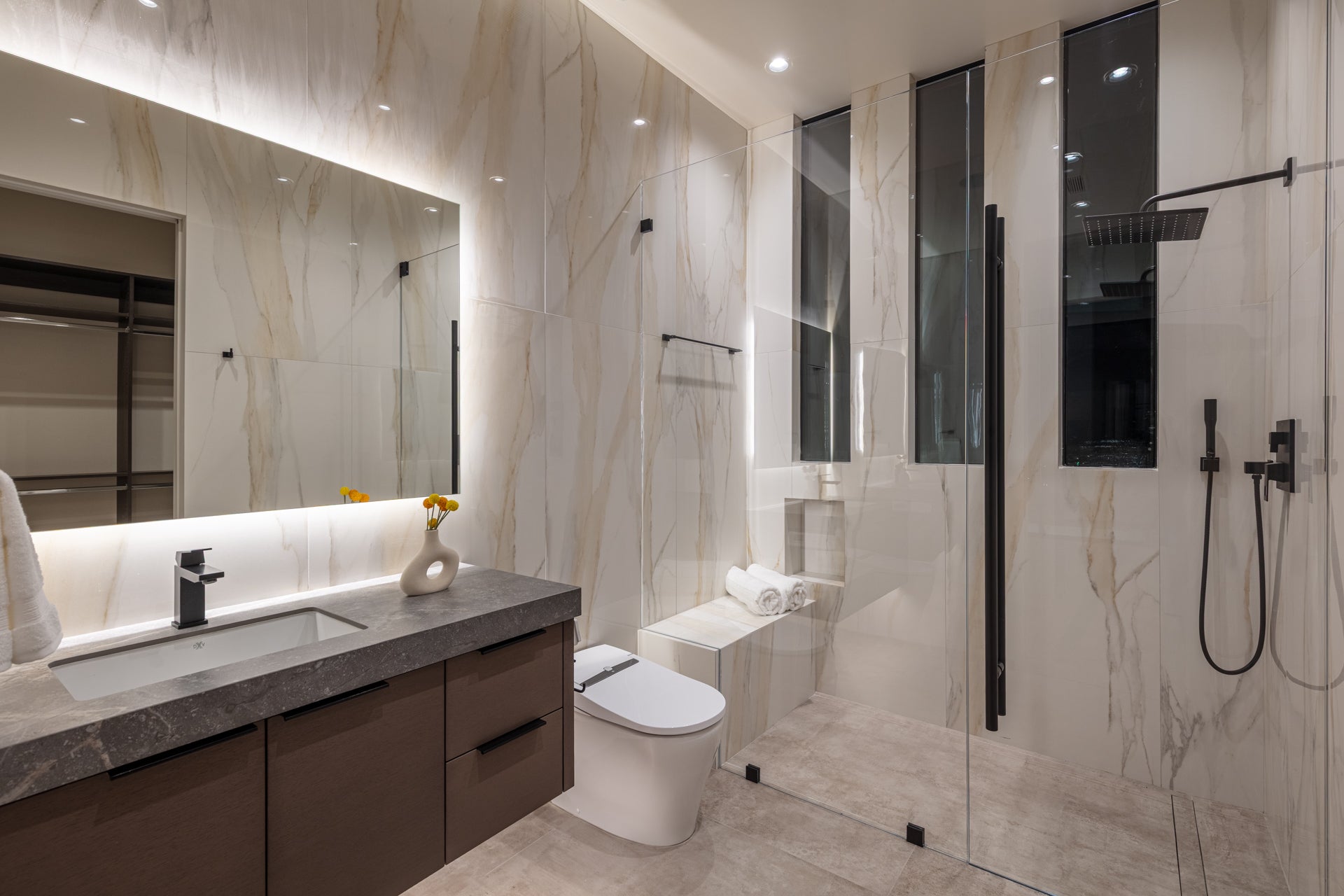 Modern bathroom with marble-look walls, a dark vanity, a back-lit mirror, and a glass shower.

