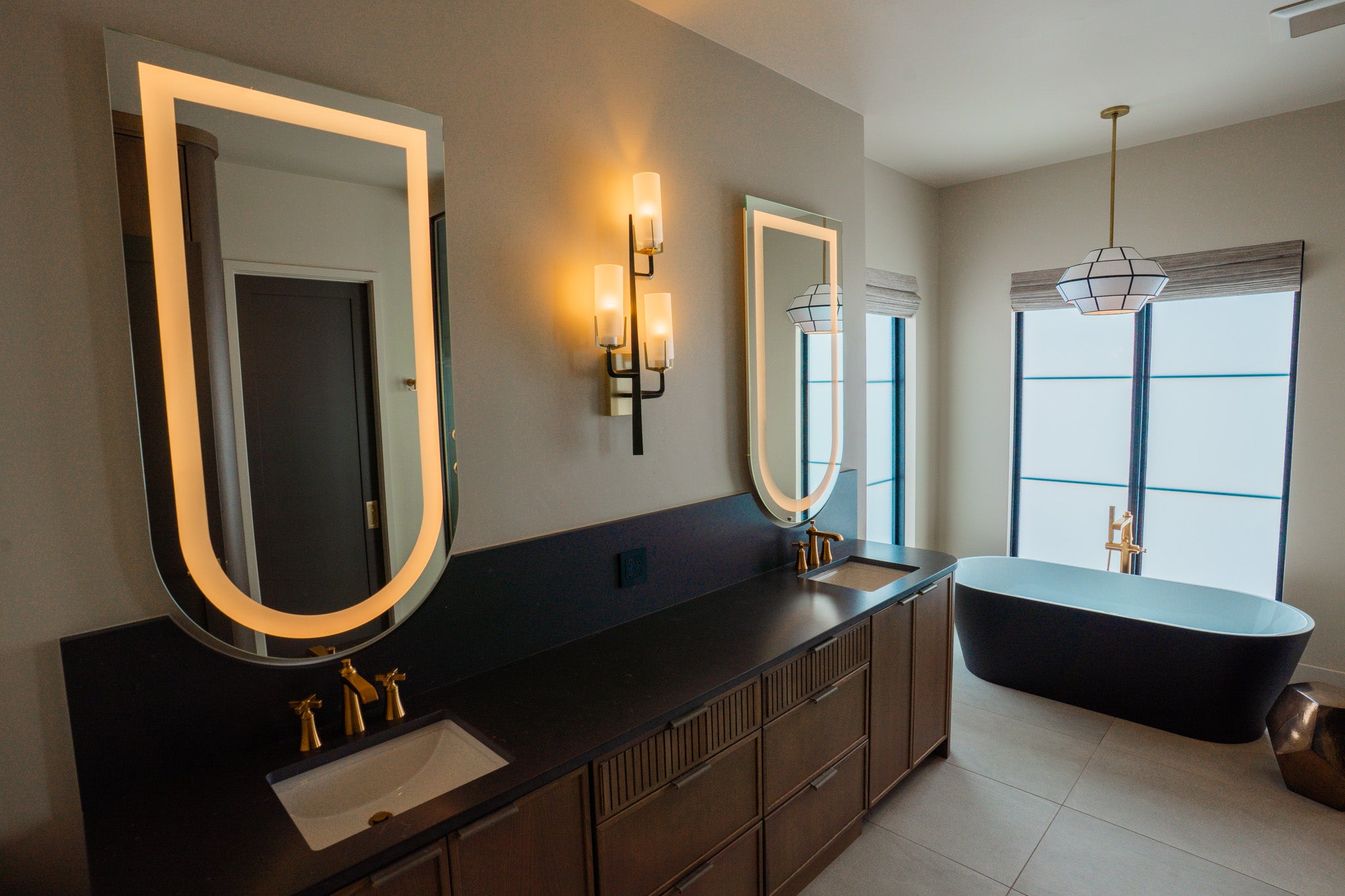 Modern bathroom with a long double vanity, two mounted soft lighted mirrors, dark cabinetry, and a freestanding tub.

