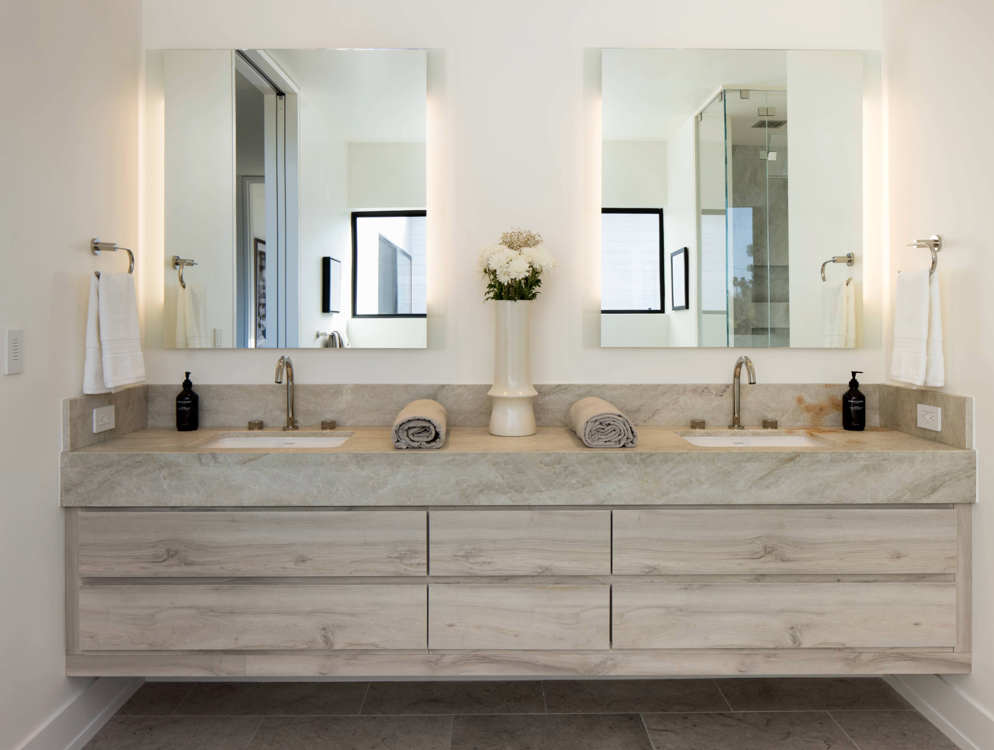 Double bathroom vanity with two wall-mounted mirrors with lights on each side, a light stone countertop, and wood cabinetry.

