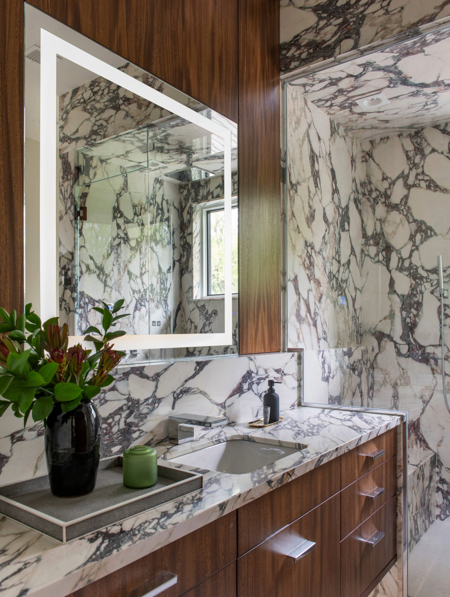 Bathroom vanity with marble-patterned surfaces, wood cabinetry, a wall-mounted mirror with task lighting, and a built-in sink.

