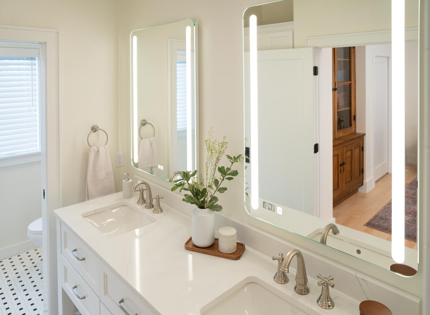 A double‑sink vanity features two rounded rectangular mirrors with vertical light strips above faucets, accented by a vase of greenery and neatly arranged toiletries.