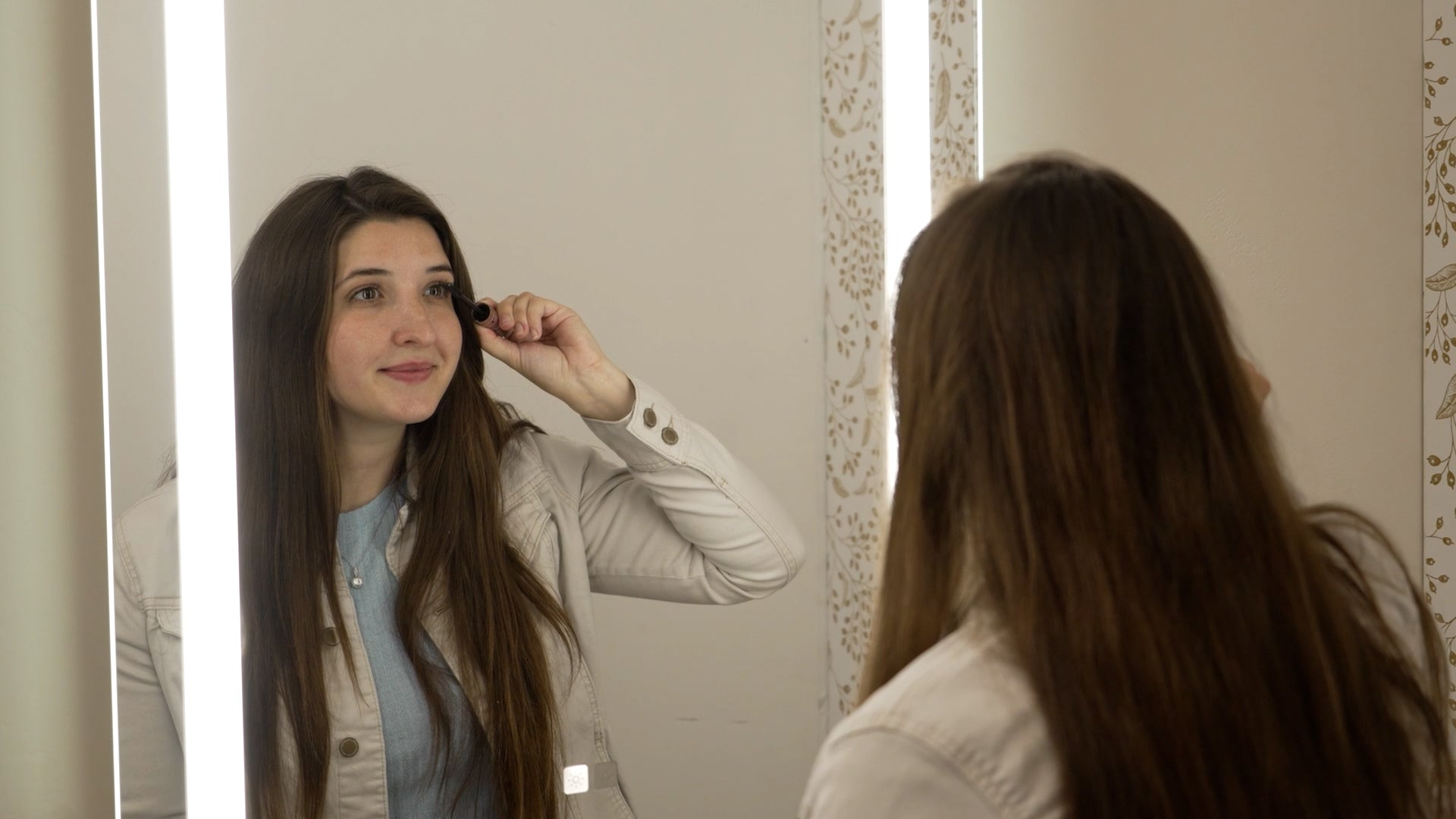 Load video: Woman applying mascara in her bathroom using a lighted mirror for better lighting.