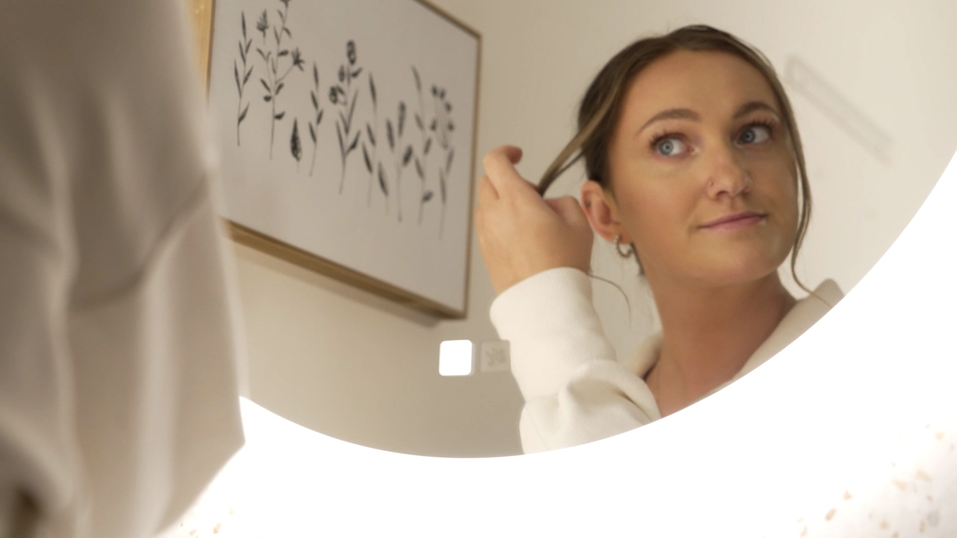 Load video: Young woman fixing hair in mirror during morning routine in her bathroom using a round lighted mirror.